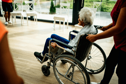 A dedicated care worker gently guiding an elderly patient in a wheelchair to a sunlit room with white chairs scattered by the window, showcasing the crucial care worker's role in providing support and mobility.