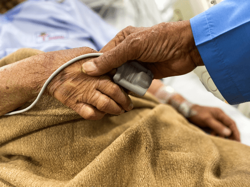 A compassionate caregiver holding the hand of an elderly patient covered by a light brown blanket, monitoring vitals with a finger machine - a symbol of attentive end-of-life care and support.
