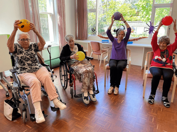 Engaged in senior care activities, four senior women sit in chairs or wheelchairs, exercising with a ball, promoting light arm movements and social connection in a room surrounded by large windows and spider decorations.