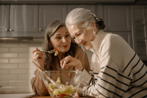 Two cheerful senior women engage in lively conversation while sharing a large bowl of salad, showcasing the essence of nutrition and dietary support in senior care.