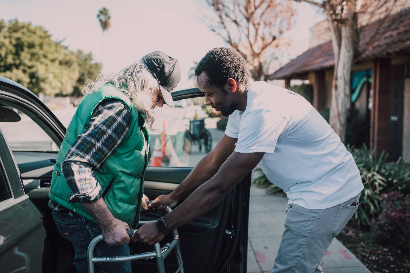 A caring young man assists a senior out of a car, holding the walker against the backdrop of a hospital, exemplifying the essence of senior care staffing.