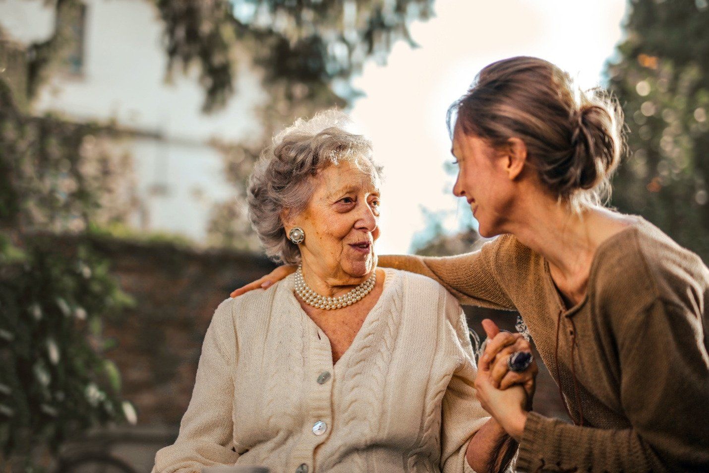 A young woman in a beige cardigan warmly greets an older woman outdoors, showcasing the joy and connection facilitated by senior care staff.