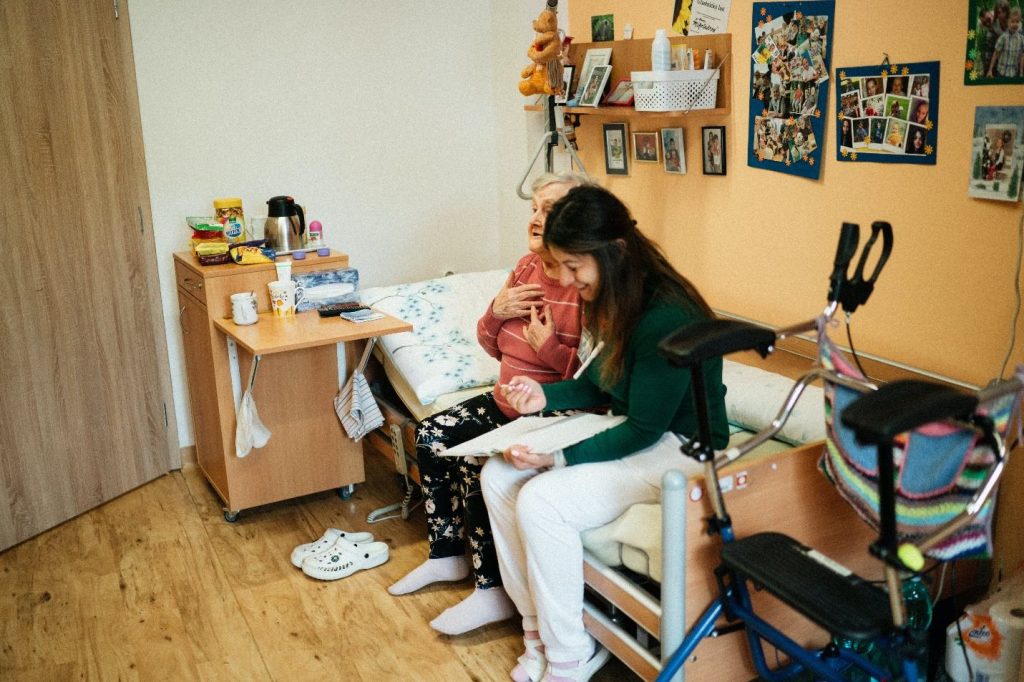 A young woman in a green jumper sits beside a senior resident at a nursing home, illustrating the caring support provided by nursing home staffing.