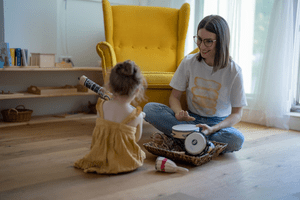 A young girl sitting on a wooden floor facing a smiling adult woman who is demonstrating a small drum.