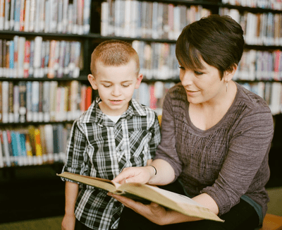 A young boy reading a book with the guidance of a caring adult woman in a library.
