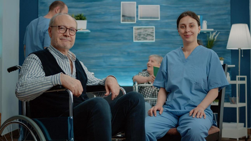 Elderly man in a wheelchair with a nurse smiling beside him at a care facility