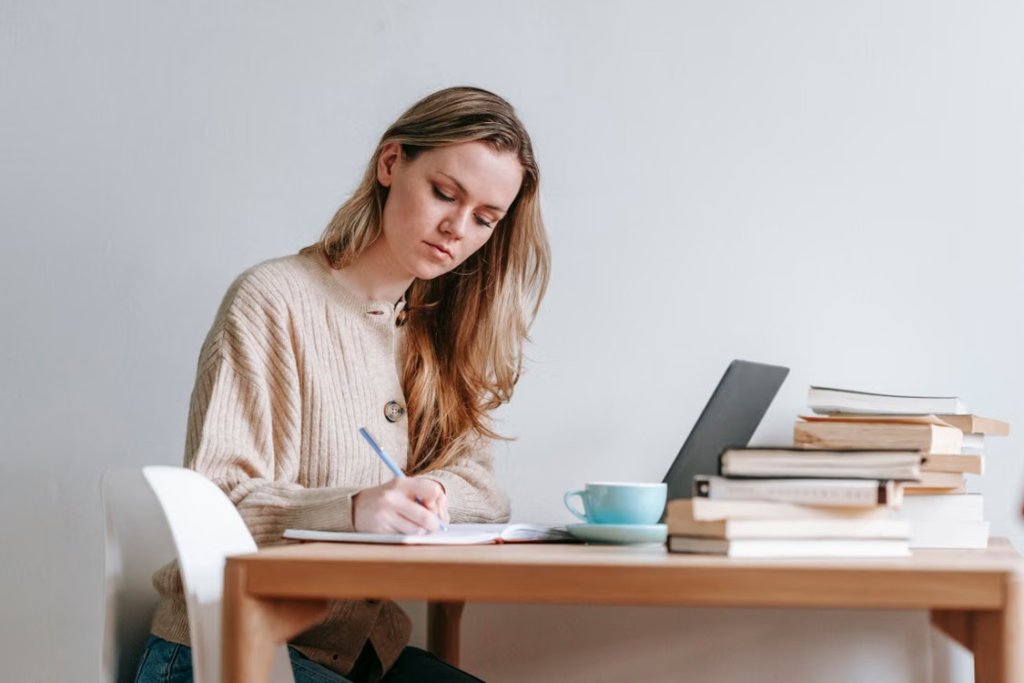 A woman writing notes at a desk with a laptop and stack of books nearby.