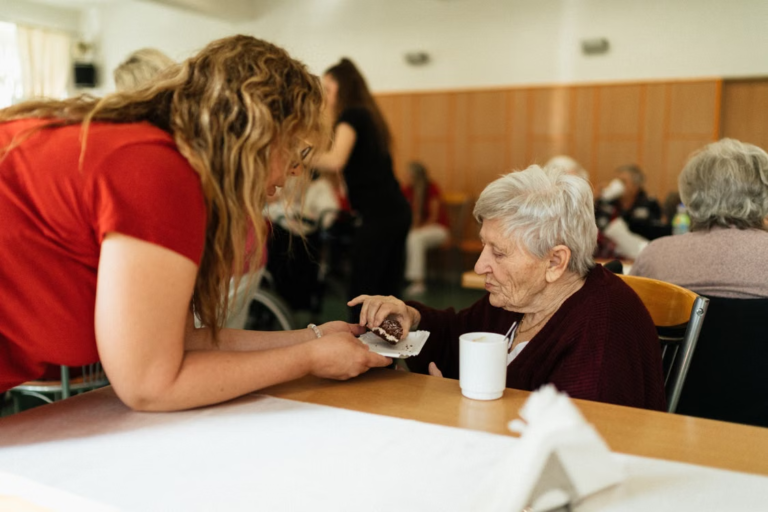 A support worker sitting with a resident, sharing tea and cake in a communal dining area.