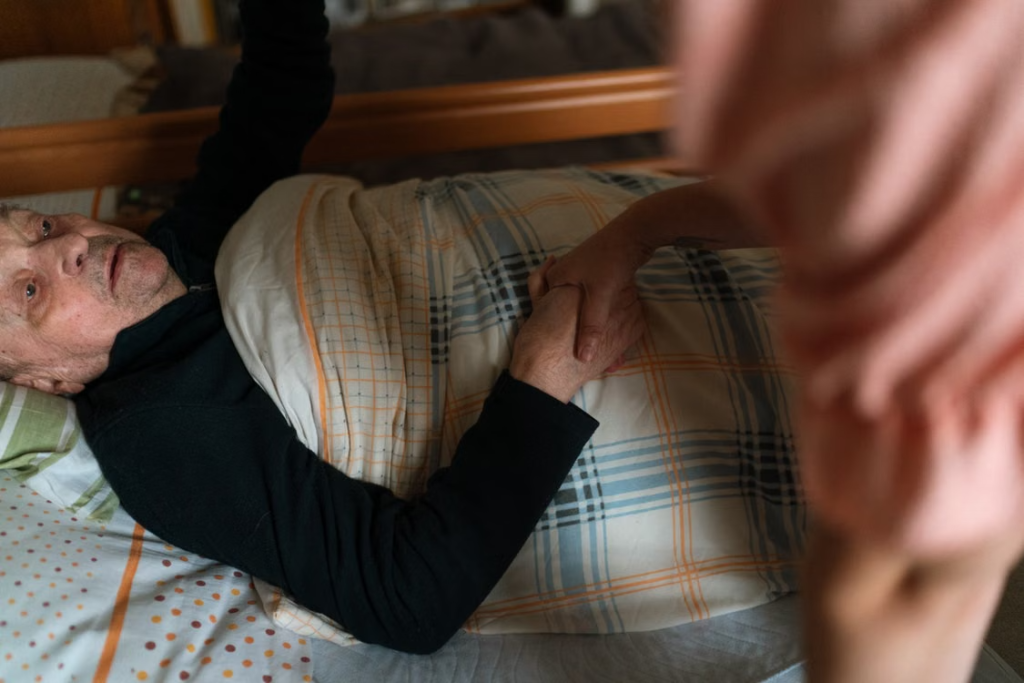 an elderly man lying on a bed holding his caregiver’s hand