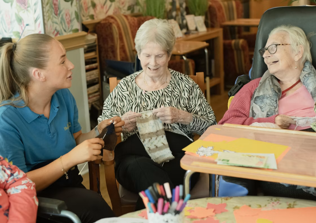a caregiver assisting two elderly women with knitting